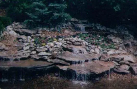 A tiered waterfall flows over rocks into a pond. Green foliage surrounds the falls.