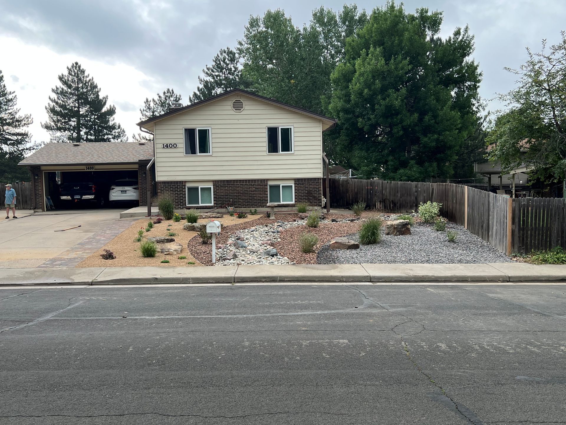 Tan house with a carport, rock garden, and a wooden fence. Gray asphalt road in front.