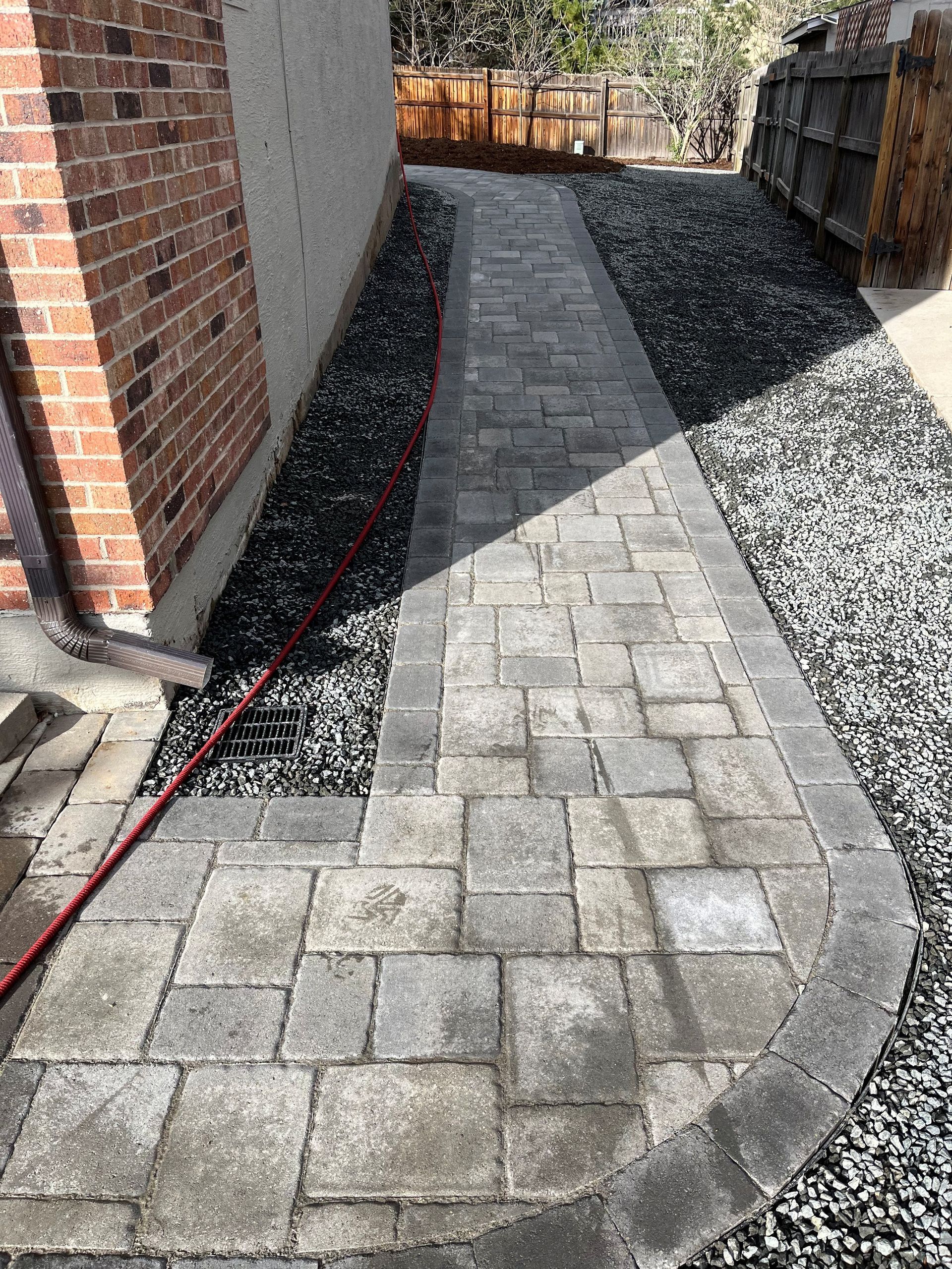 Brick paver walkway next to a brick wall and gravel, leading toward a wooden fence.