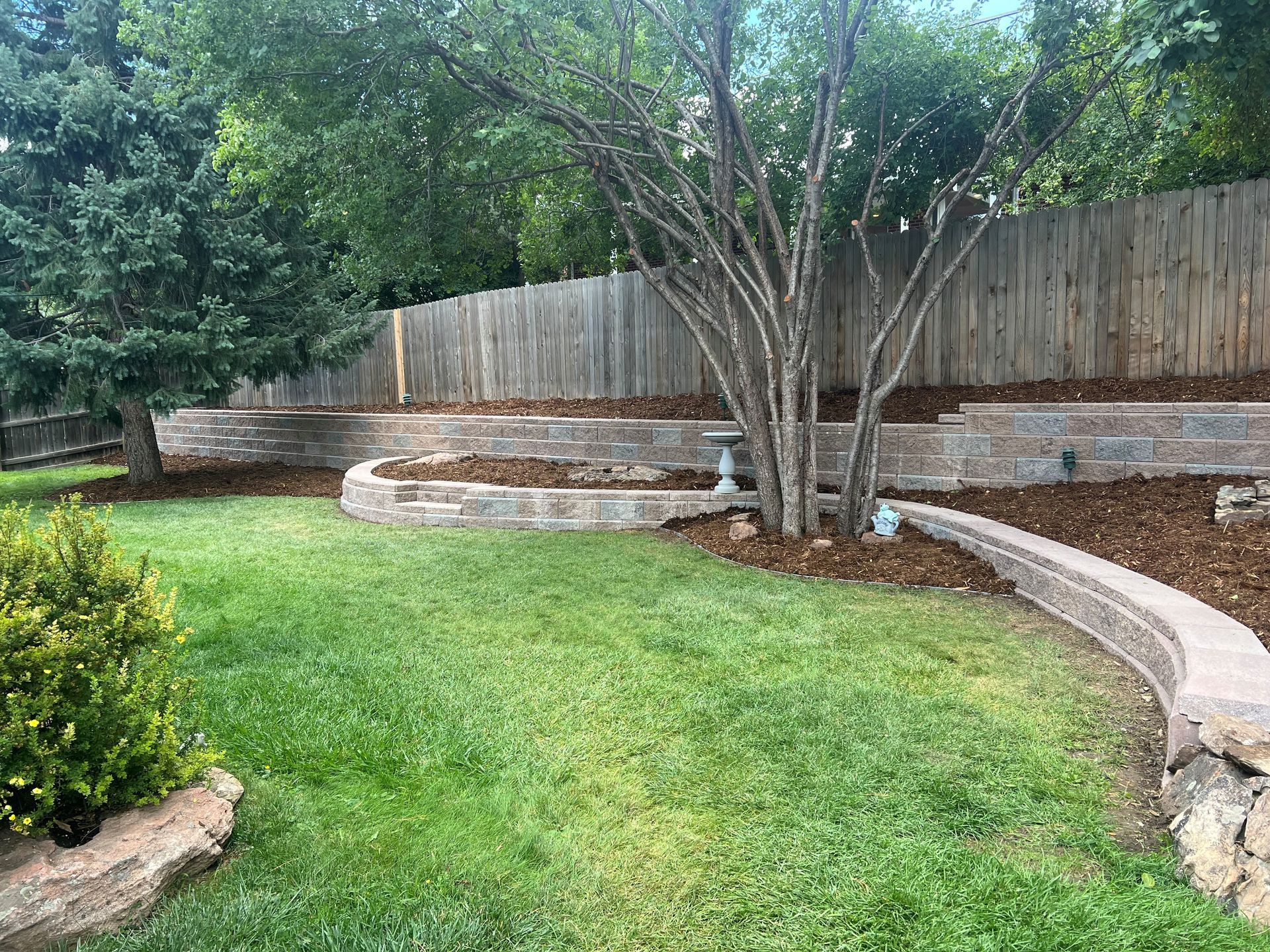 Backyard with tiered retaining walls, green grass, and trees against a wooden fence.