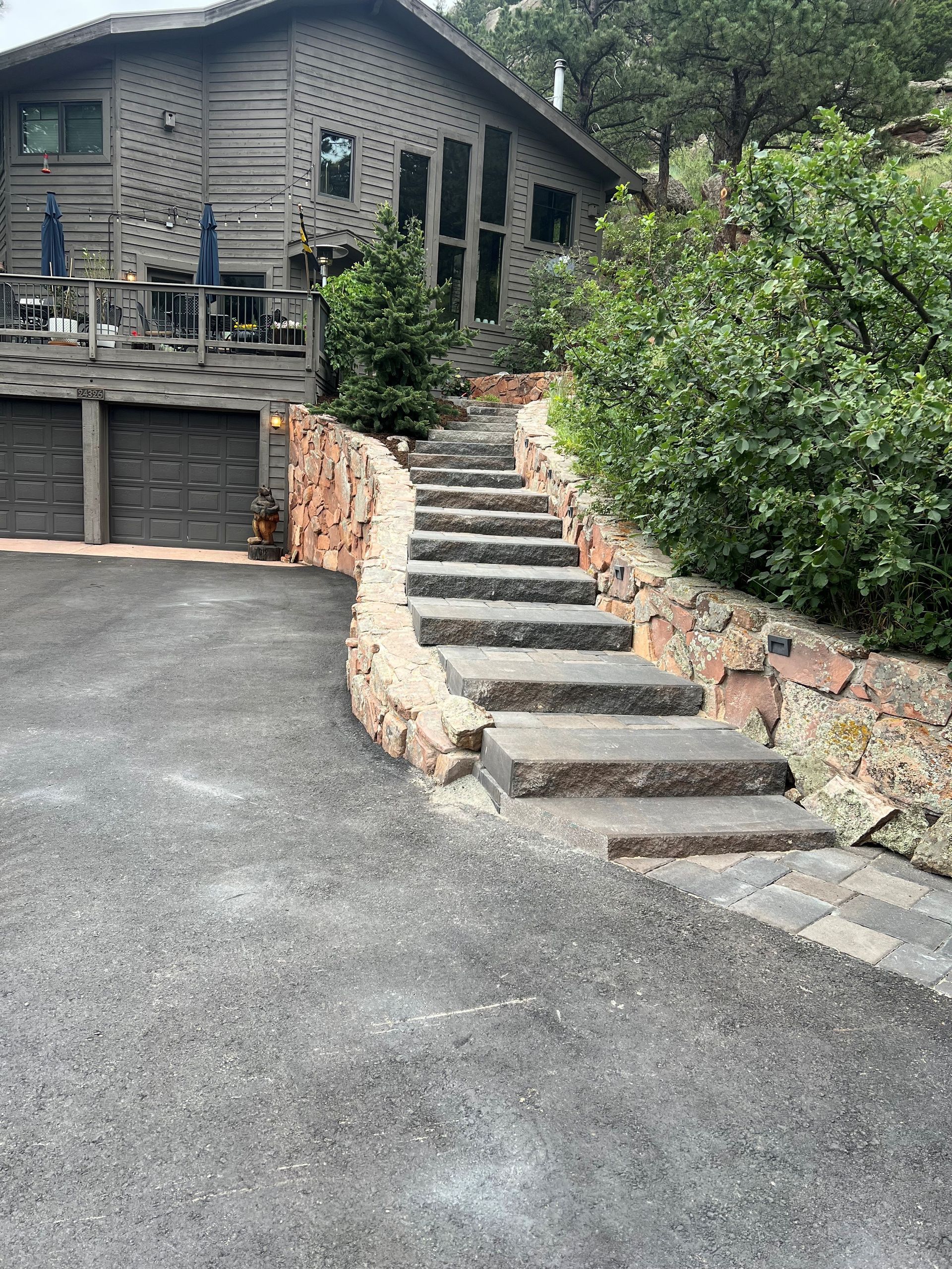 Stone steps leading up to a wooden house with a paved driveway and surrounding greenery.