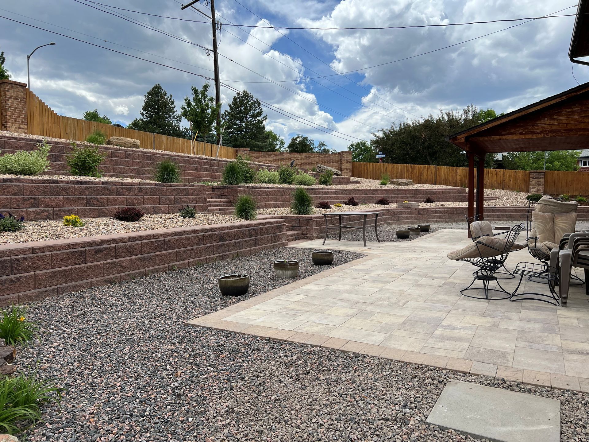 A tiered backyard with retaining walls, patio, gravel, and plants under a cloudy sky.