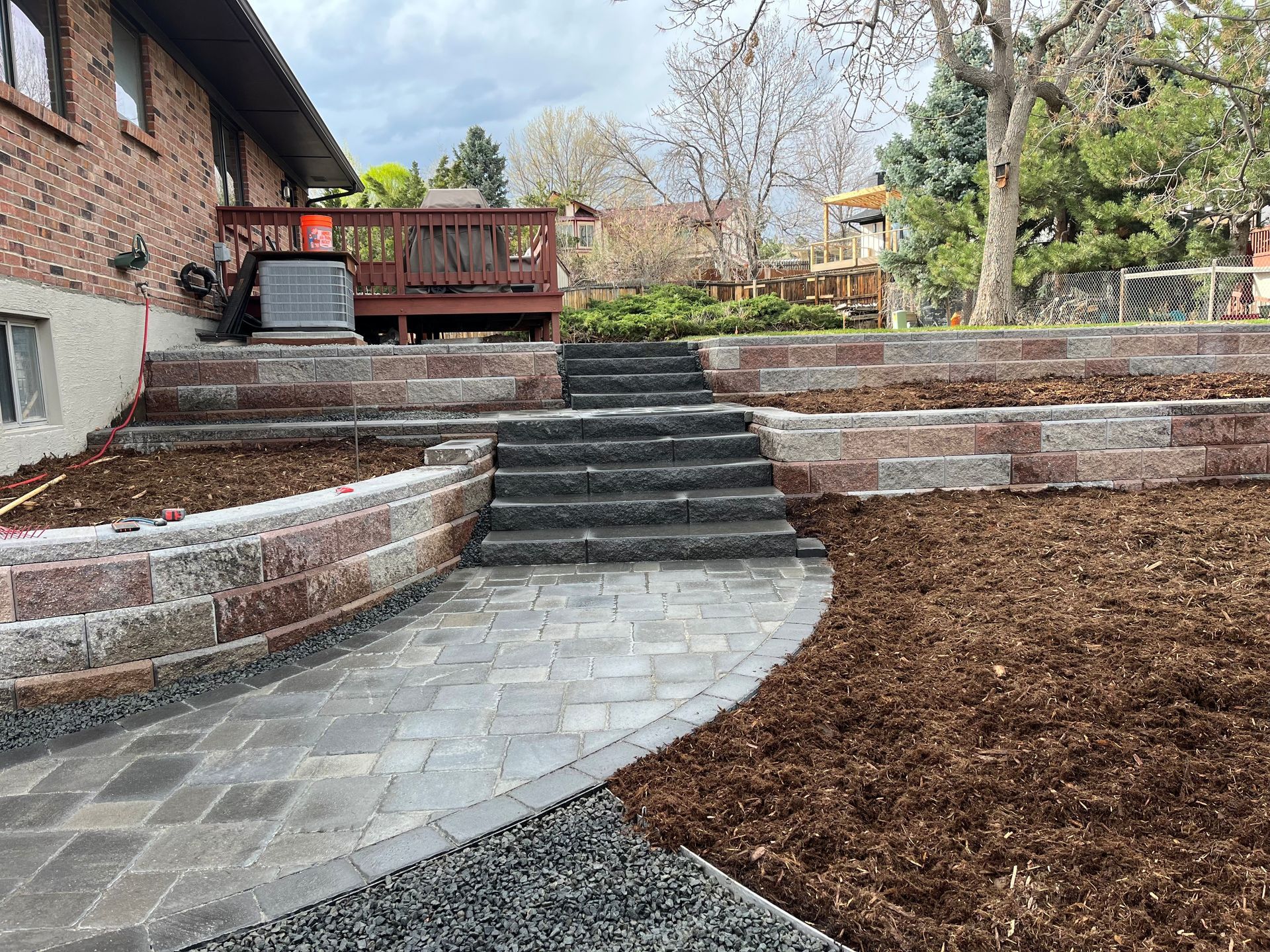 Brick retaining walls and steps leading to a wooden deck in a backyard, with a paved path and mulch.