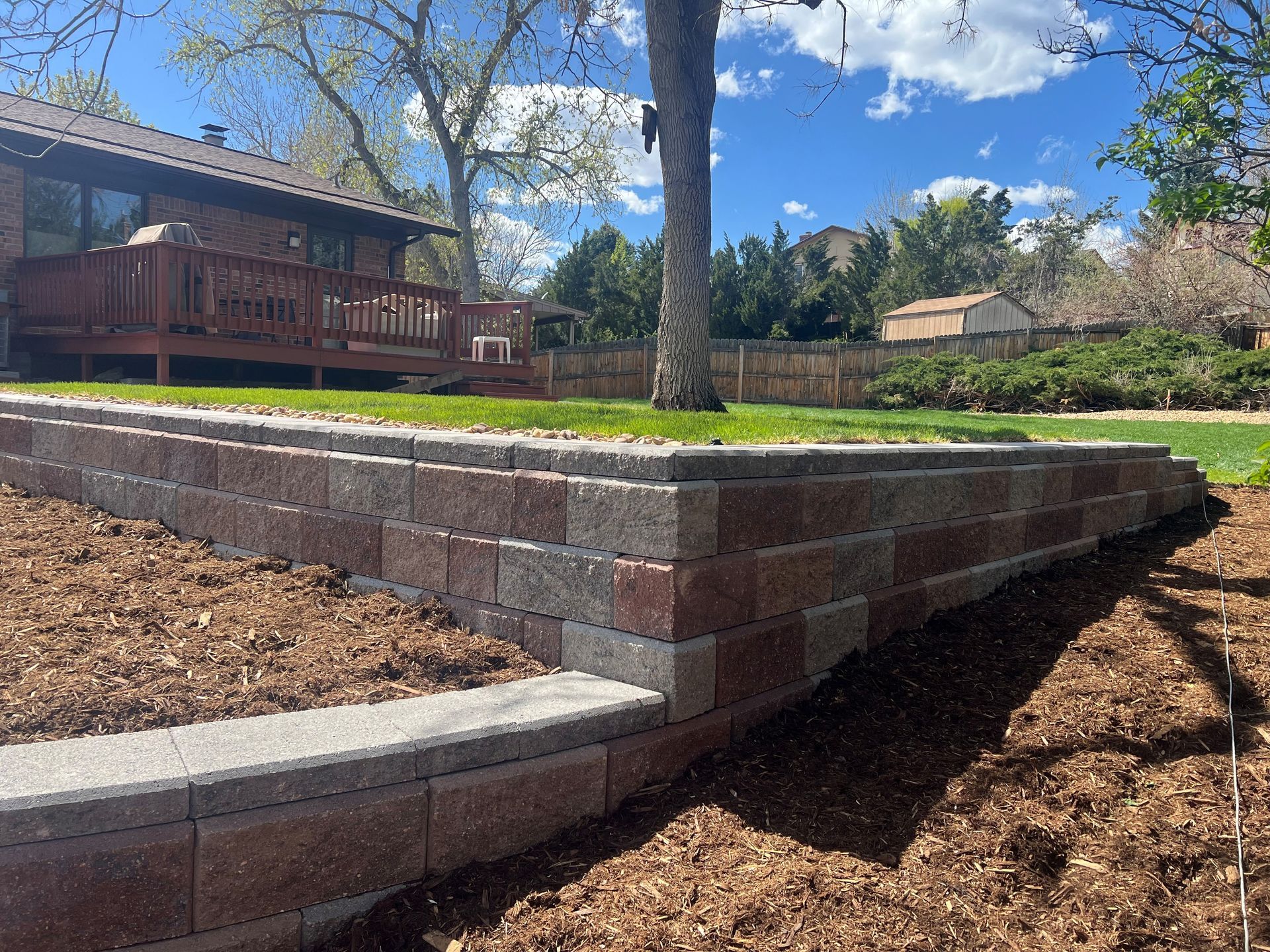 Brick retaining wall in yard, brown and gray bricks, topped with grass, tree in background, sunny day.