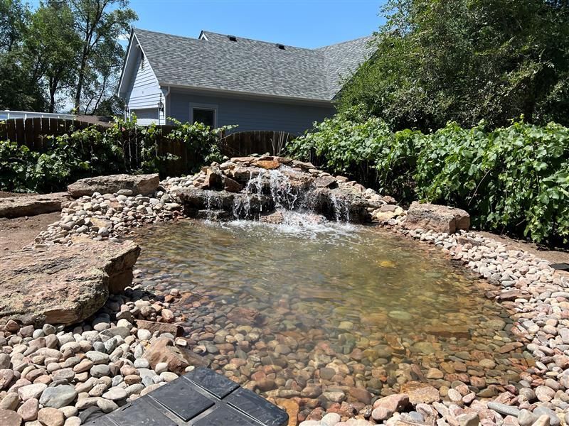 Backyard pond with small waterfall, surrounded by rocks and gravel, in front of a house.