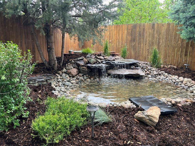 Pond with small waterfall and stepping stones surrounded by rocks and plants in a backyard.