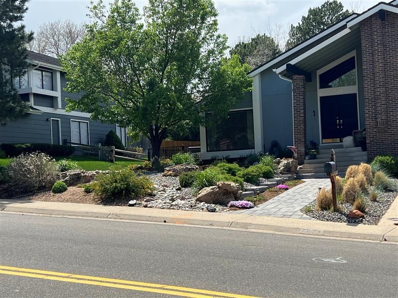 House with landscaped front yard, including rocks, plants, and a brick path. Another house visible in the background.