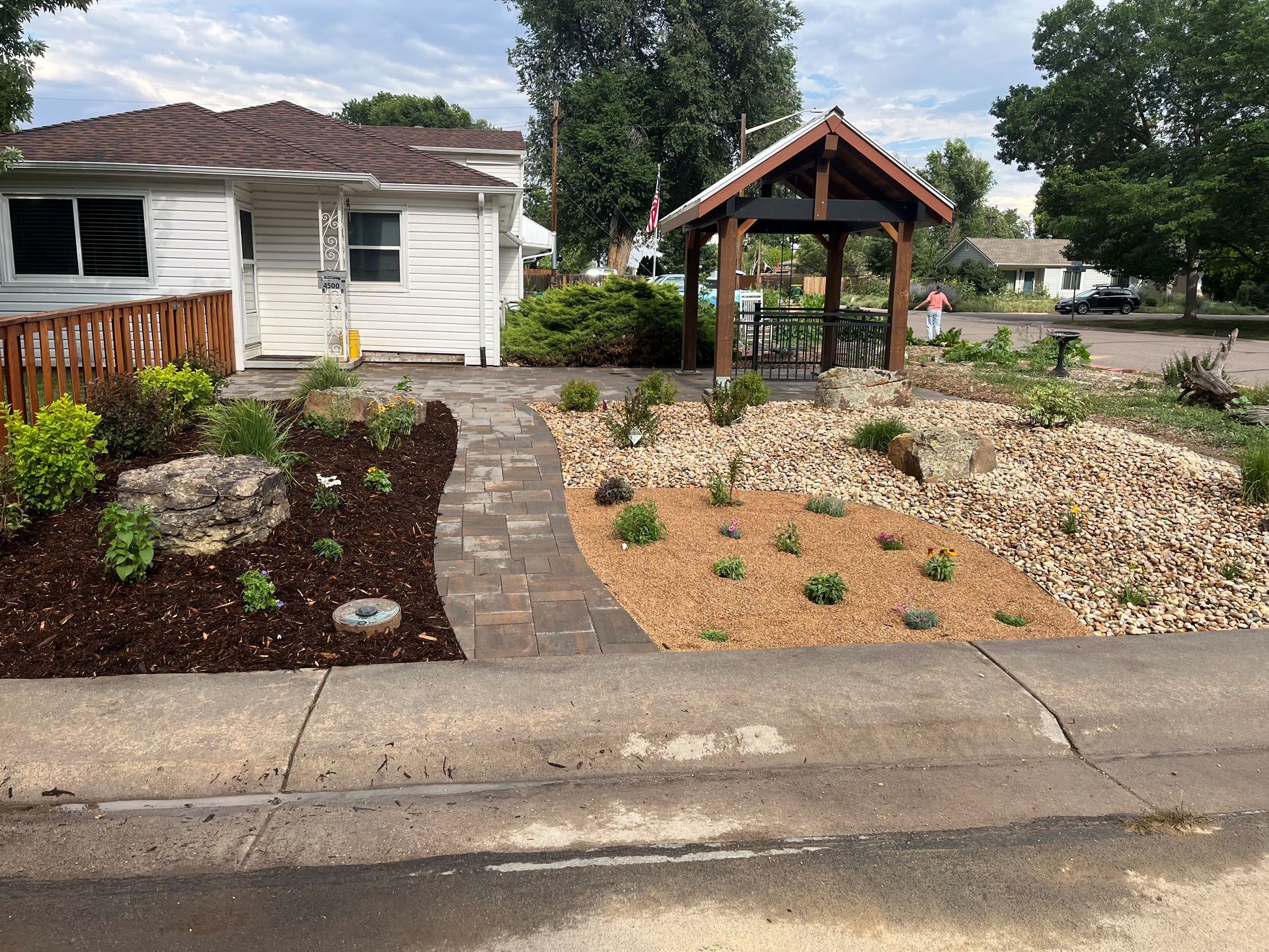 A small white house with a landscaped front yard with a brick pathway and a wooden gazebo.