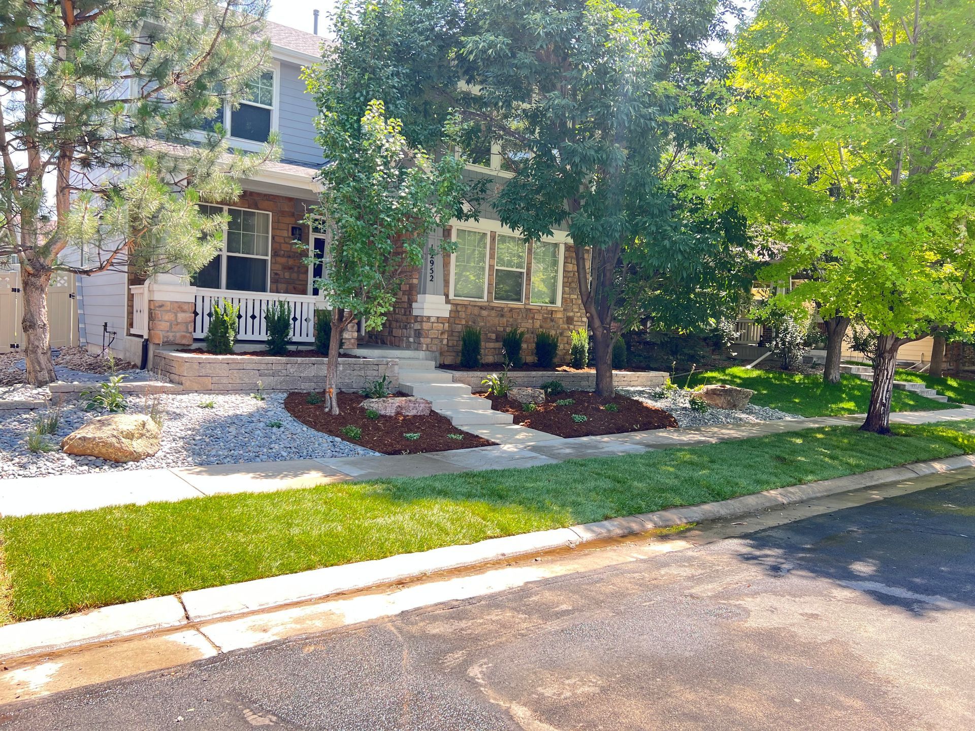 Two-story house with stone and siding facade, landscaping with trees, lawn, and rock beds. Sunny day.