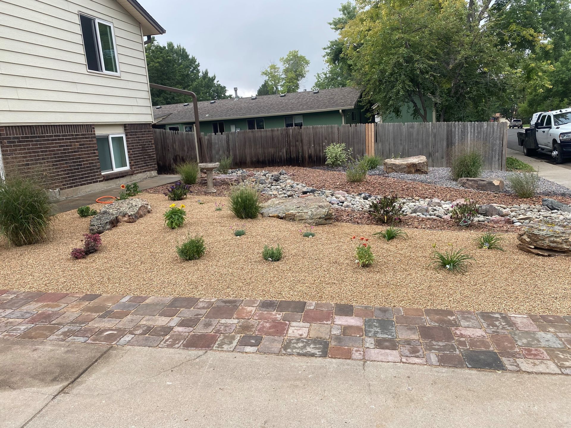 Rock garden with tan gravel, scattered plants, and brick border in front of a house.