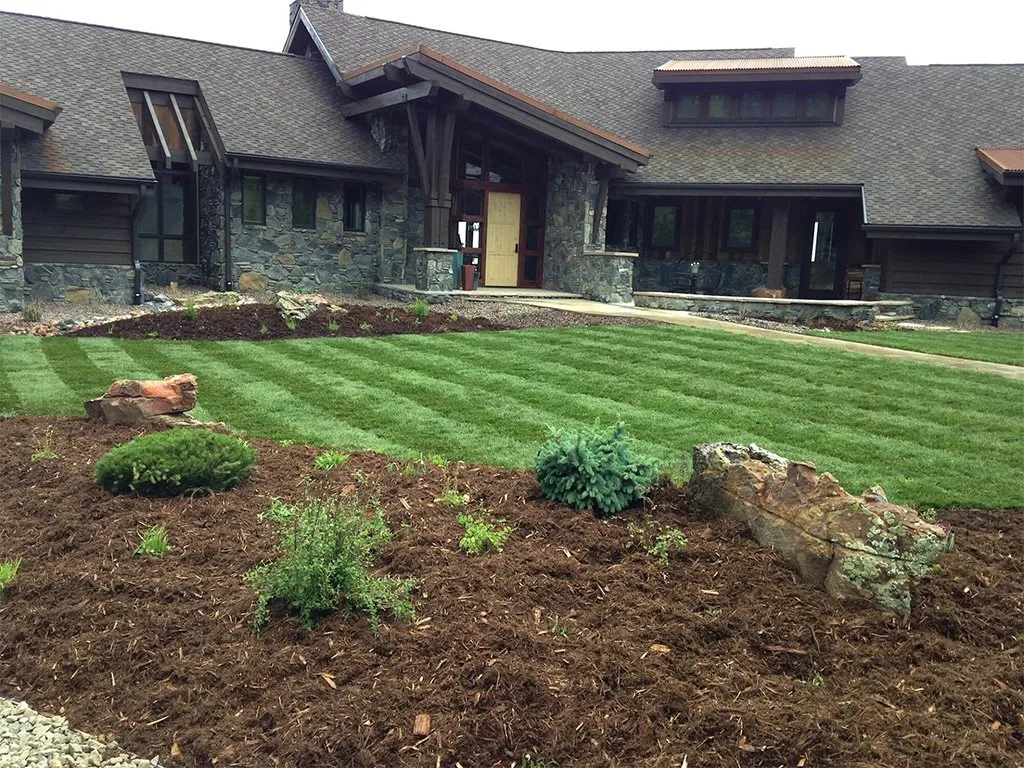 Lush green lawn with mulch and plants in front of a large brown stone house.
