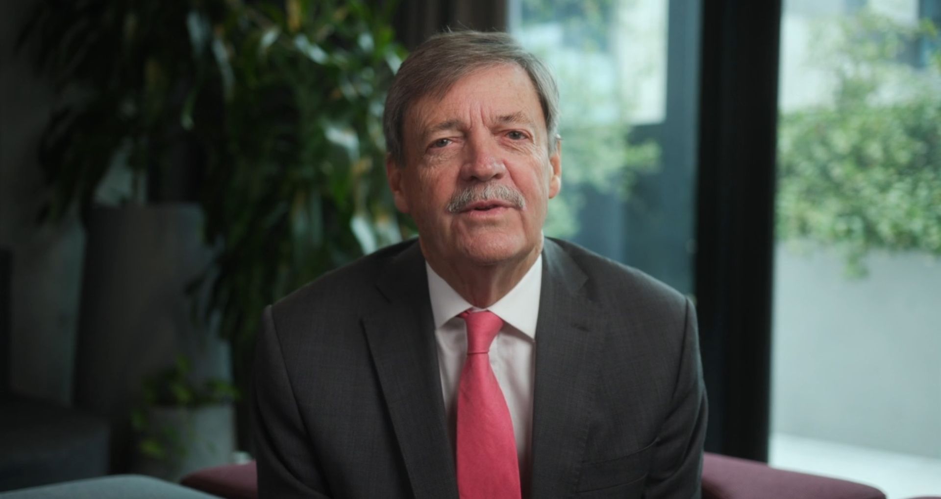 Man in a dark suit and pink tie seated indoors, speaking with a neutral expression by a window