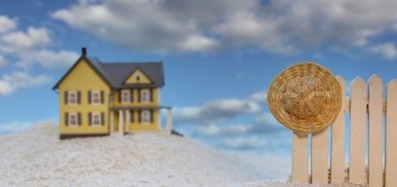 Miniature yellow house on snowy ground, with a wicker ornament and pale fence under a blue sky.