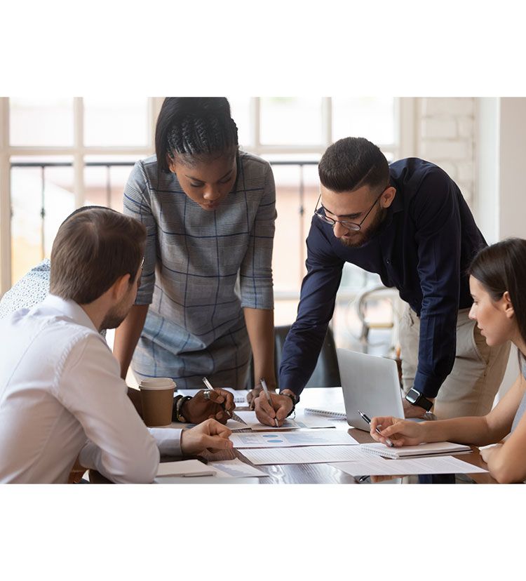 Four coworkers leaning over a table, discussing documents and a laptop in a bright office meeting room