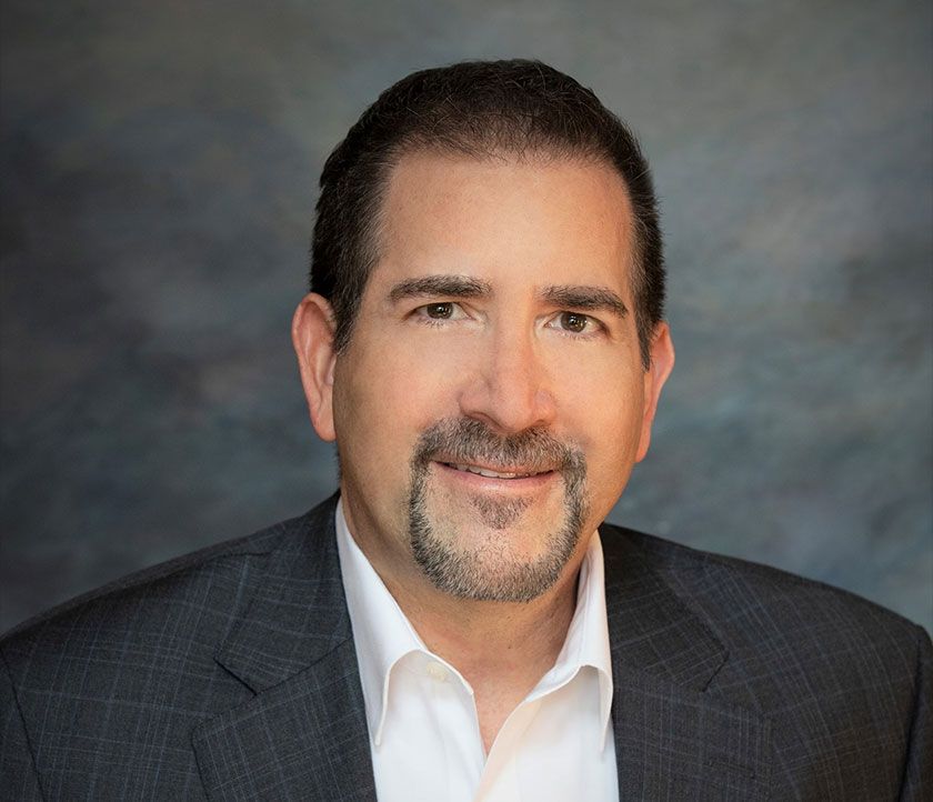 Studio headshot of a man in a dark blazer and white shirt, smiling against a gray backdrop
