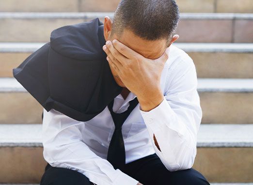 Person sitting on steps, covering their face with one hand and holding a black jacket over one shoulder.
