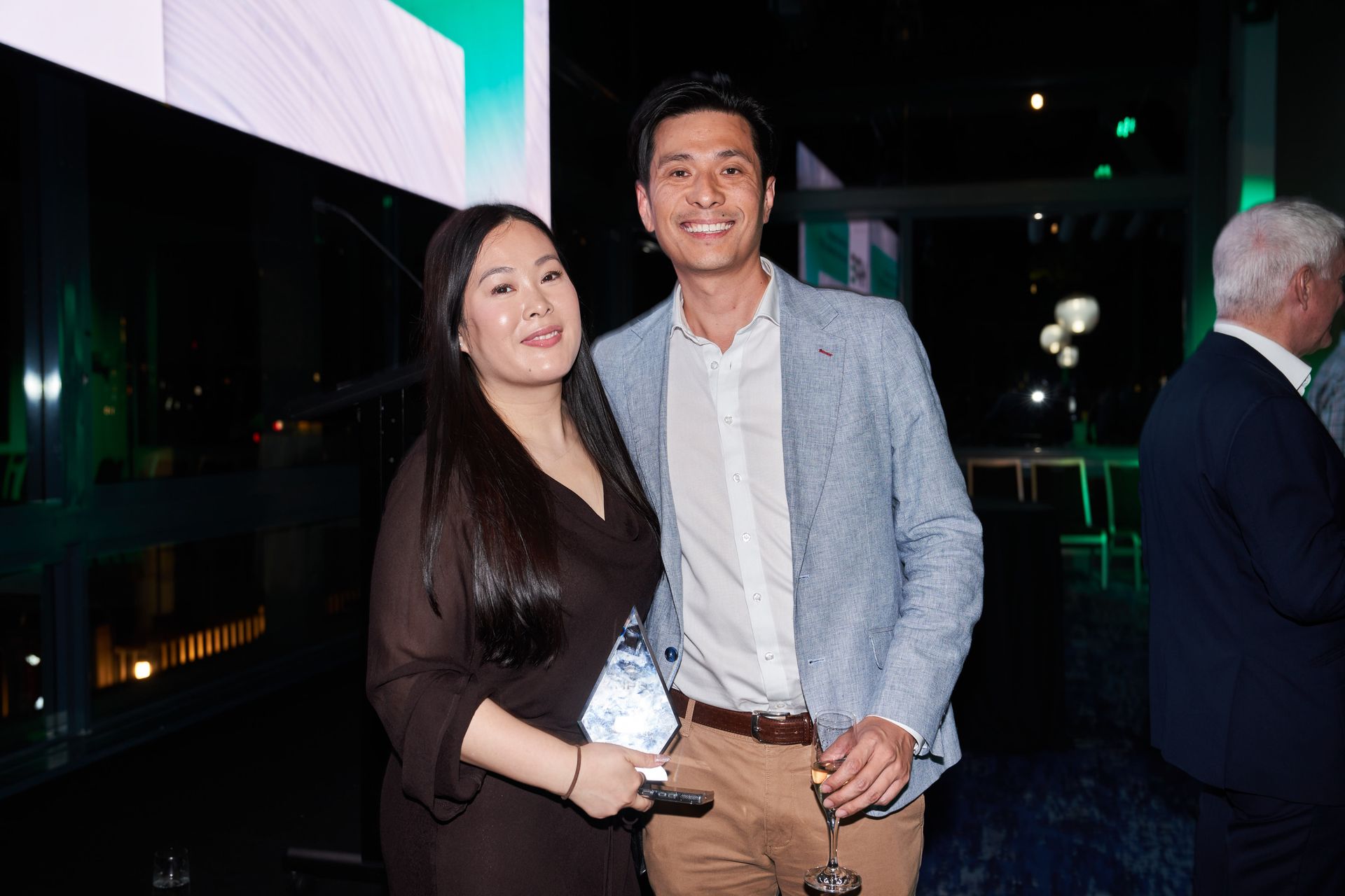 Two people smiling, holding an award. Woman in black dress, man in blue jacket, both at an event.
