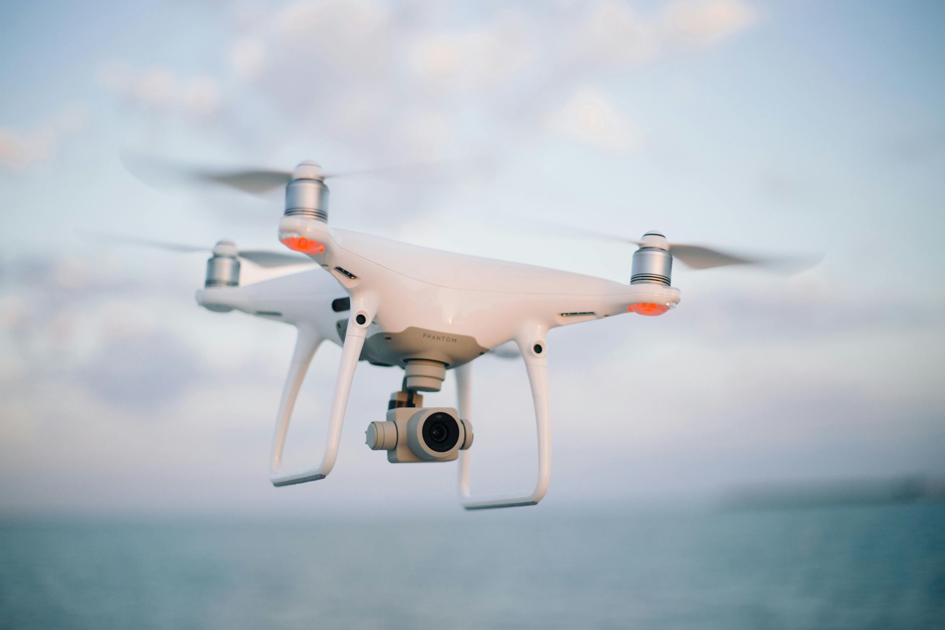 White drone with spinning propellers hovers in mid-air, blue water and sky in the background.