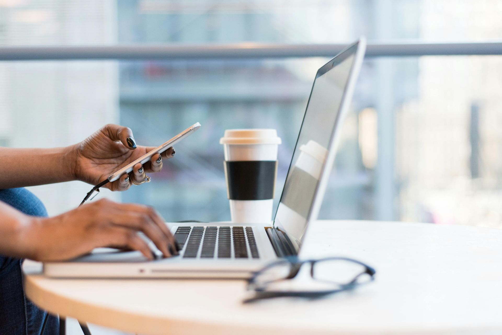 Person working on laptop, holding phone, coffee cup and glasses on table in a modern setting.