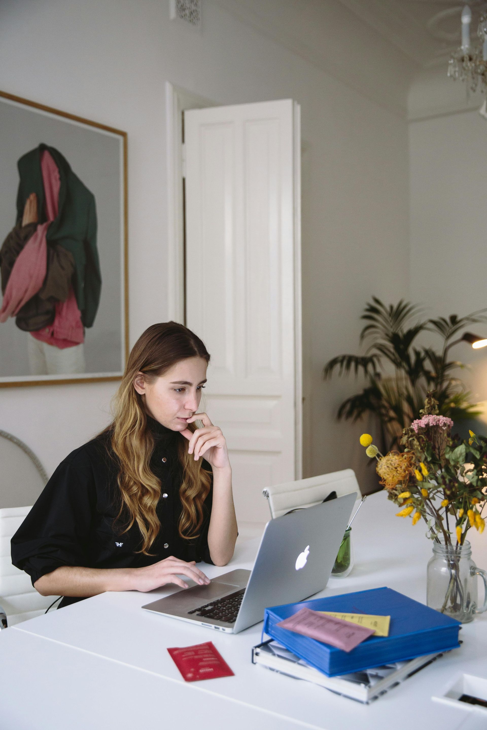 Woman with curly hair, wearing a black sweater, thoughtfully working on a laptop at a white table.