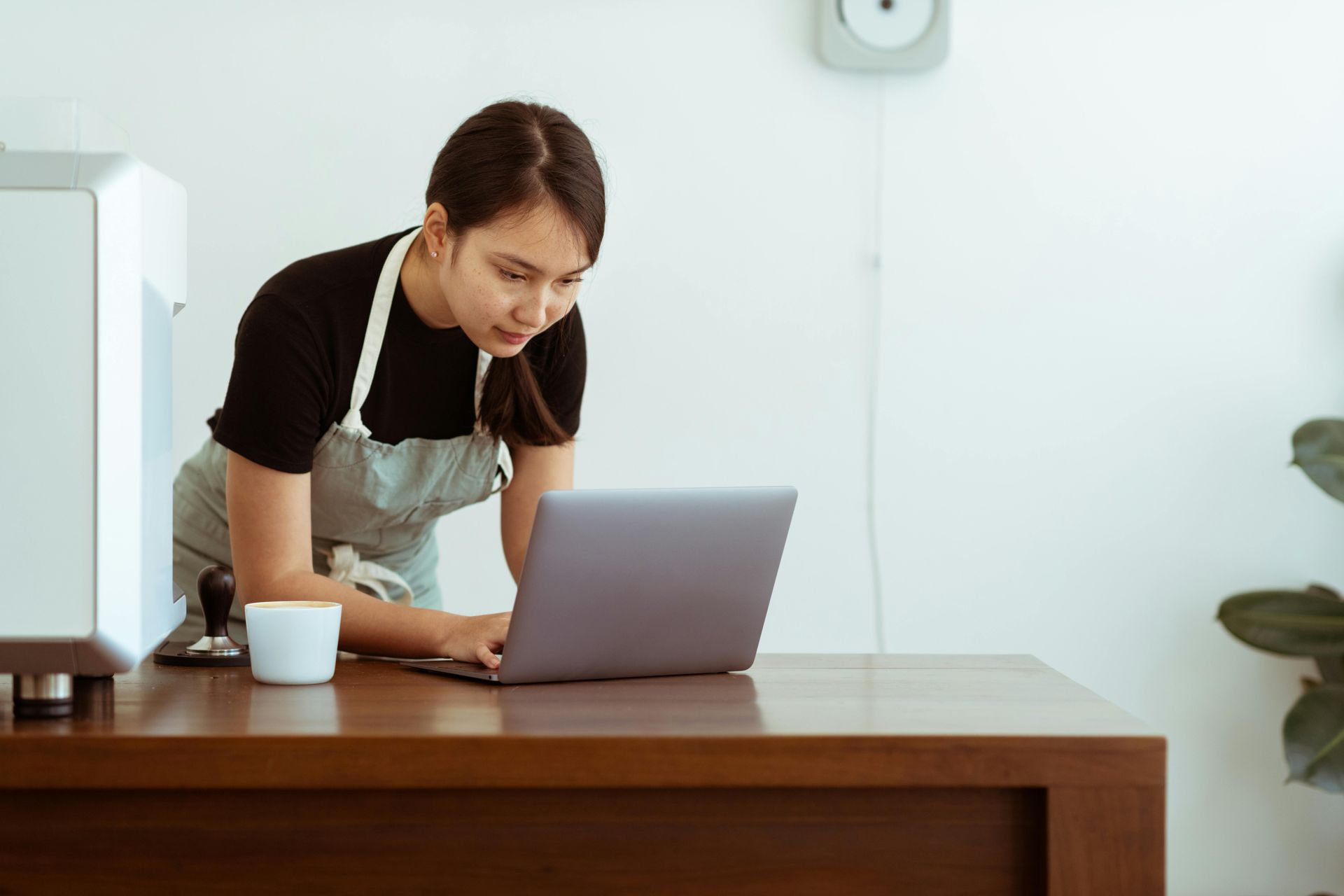 Woman wearing an apron and looking at a laptop while leaning on a wooden counter in a coffee shop.