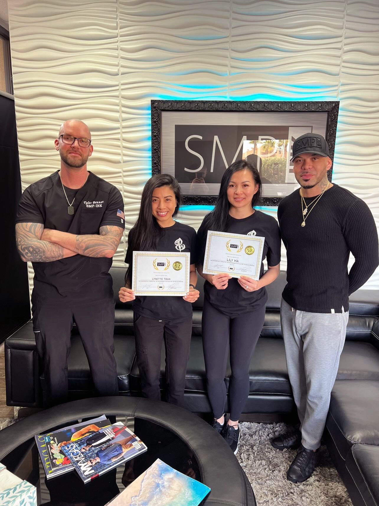 Four people posing indoors, two holding certificates, in front of a textured wall and framed sign.