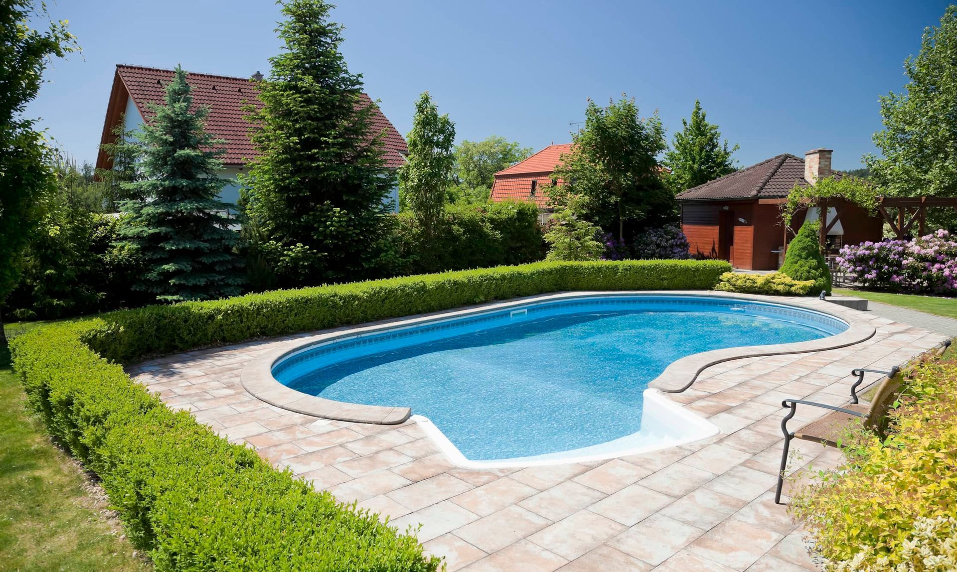Swimming pool surrounded by manicured hedges and brick patio, with houses in the background under a blue sky.