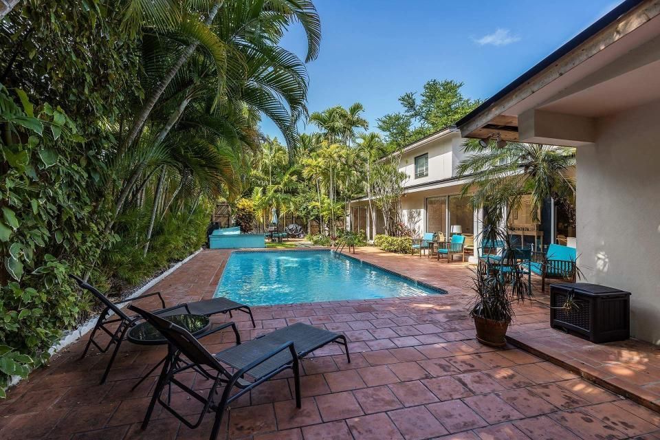 Backyard with pool, patio, lounge chairs, and two-story house surrounded by lush greenery under a blue sky.