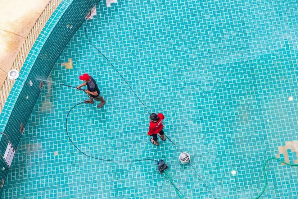 Two workers in a turquoise pool, cleaning with hoses.