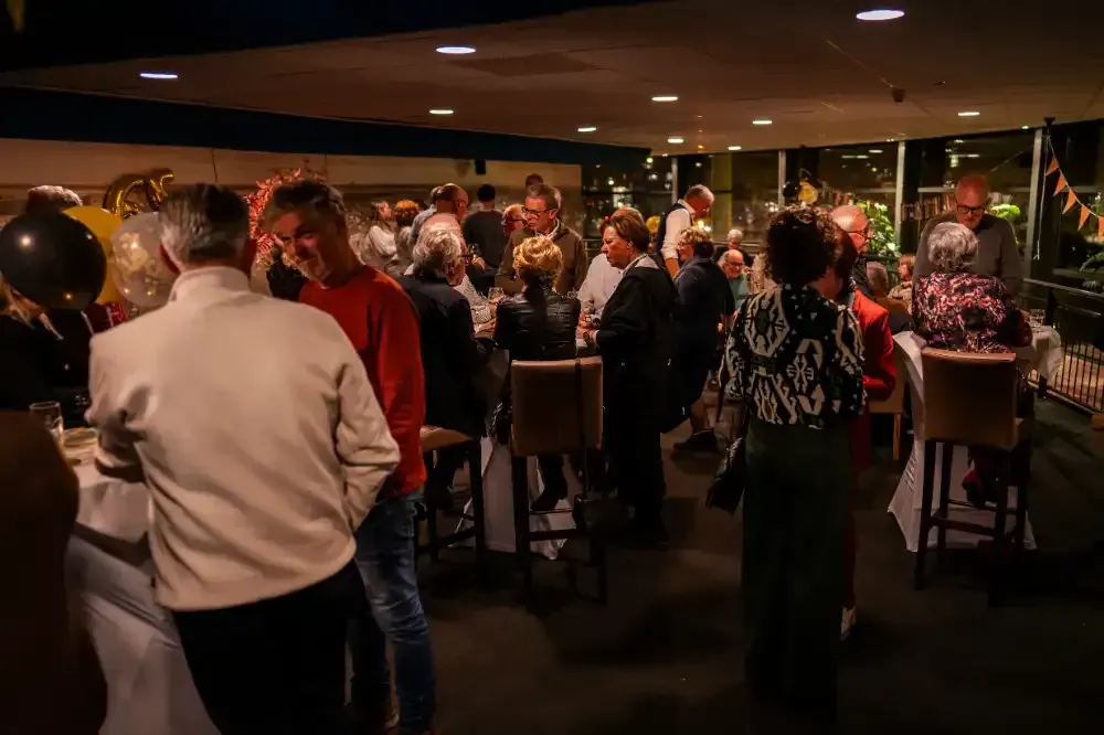 een groep mensen staat op een feestje in een kamer rond tafels.