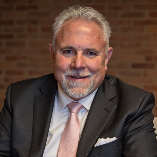 Smiling man in a dark suit and pink tie seated before a brick wall