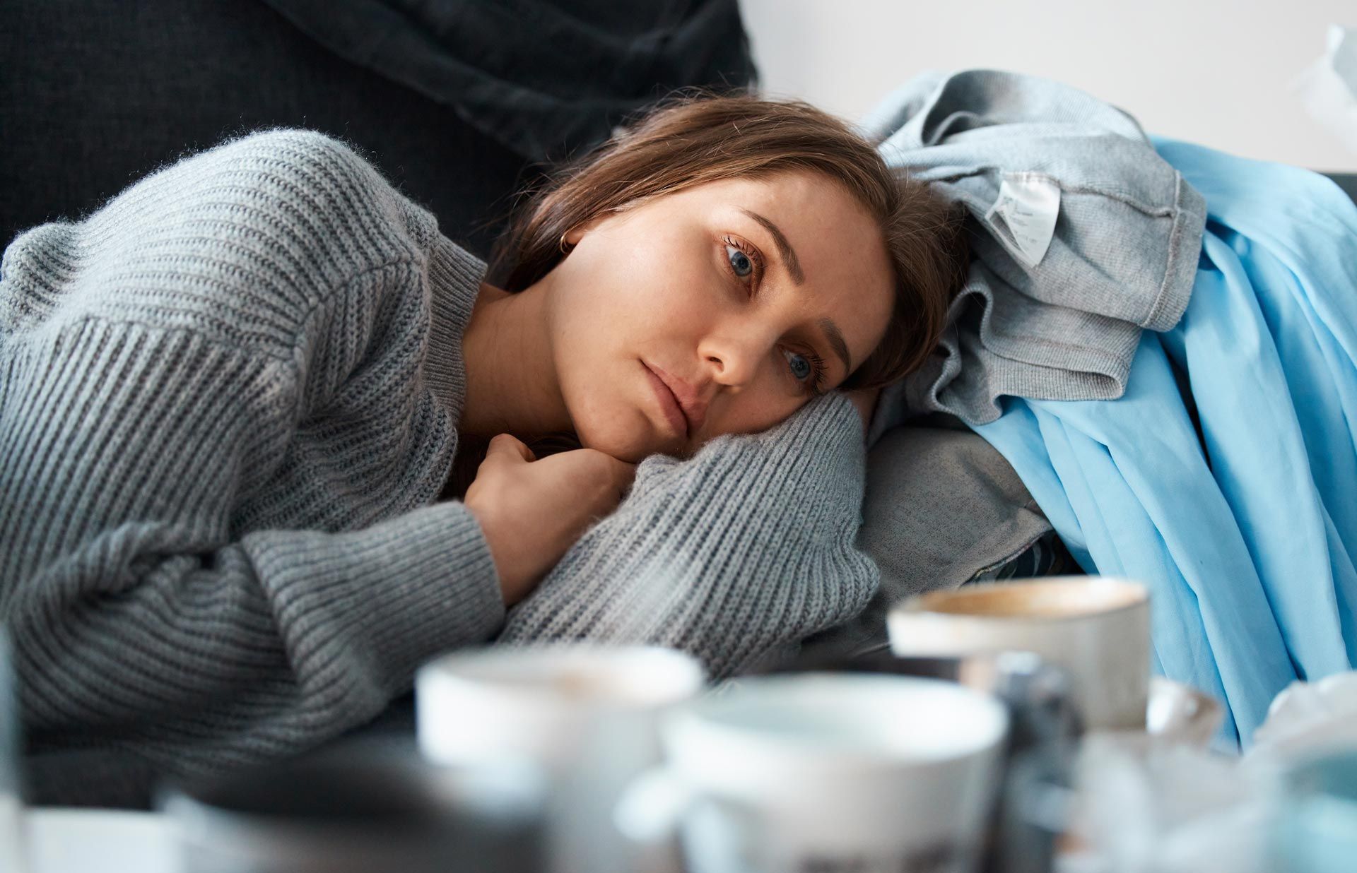 Woman resting on a couch in a gray sweater, looking tired beside a blue blanket and cups on a table