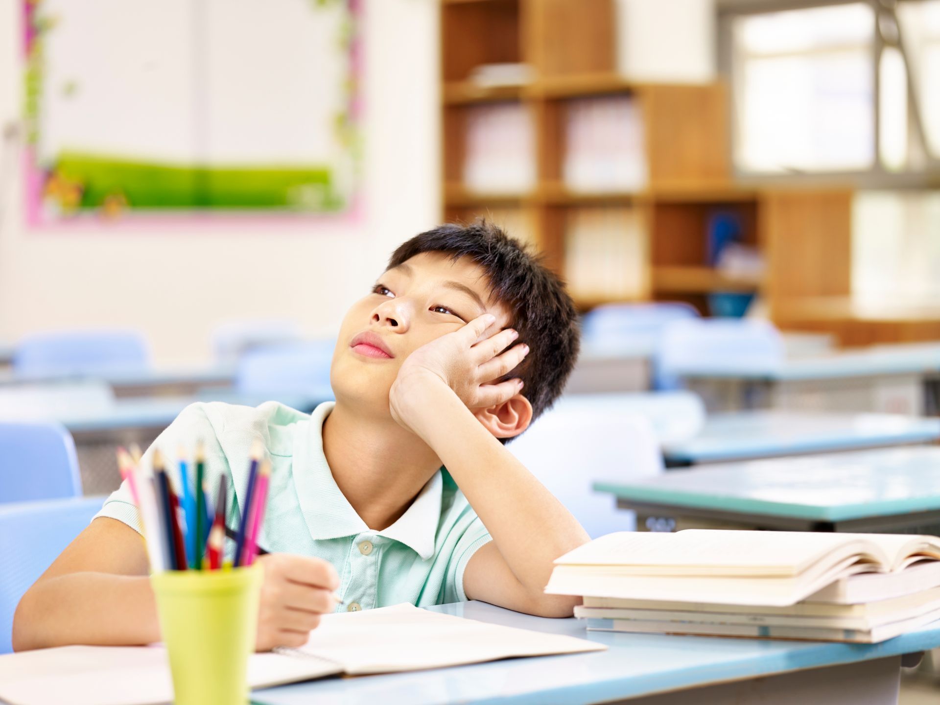 Student resting head on hand at a classroom desk with books and colorful pencils nearby