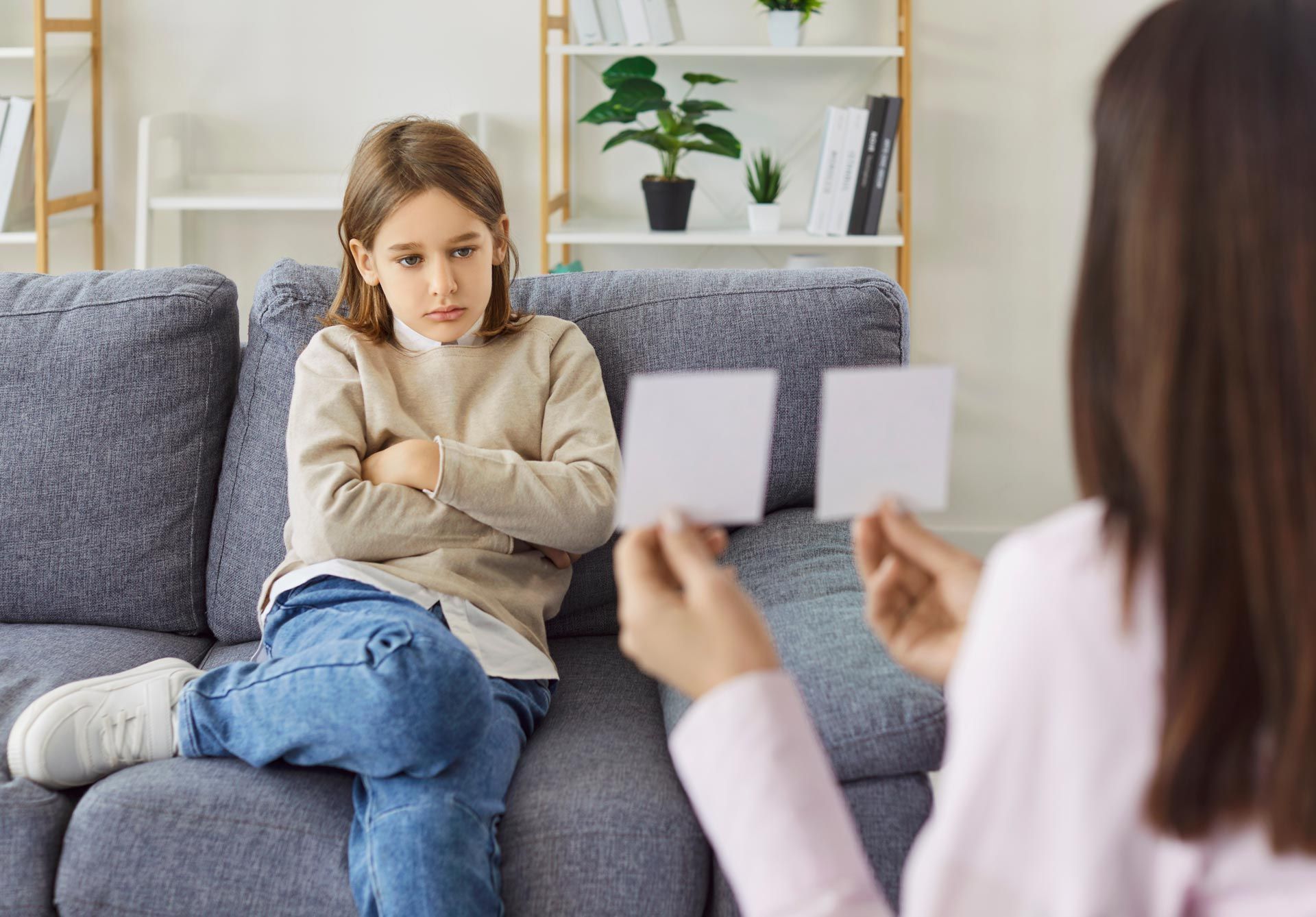 Child sitting on a gray couch, arms crossed, talking to a counselor holding a note card.