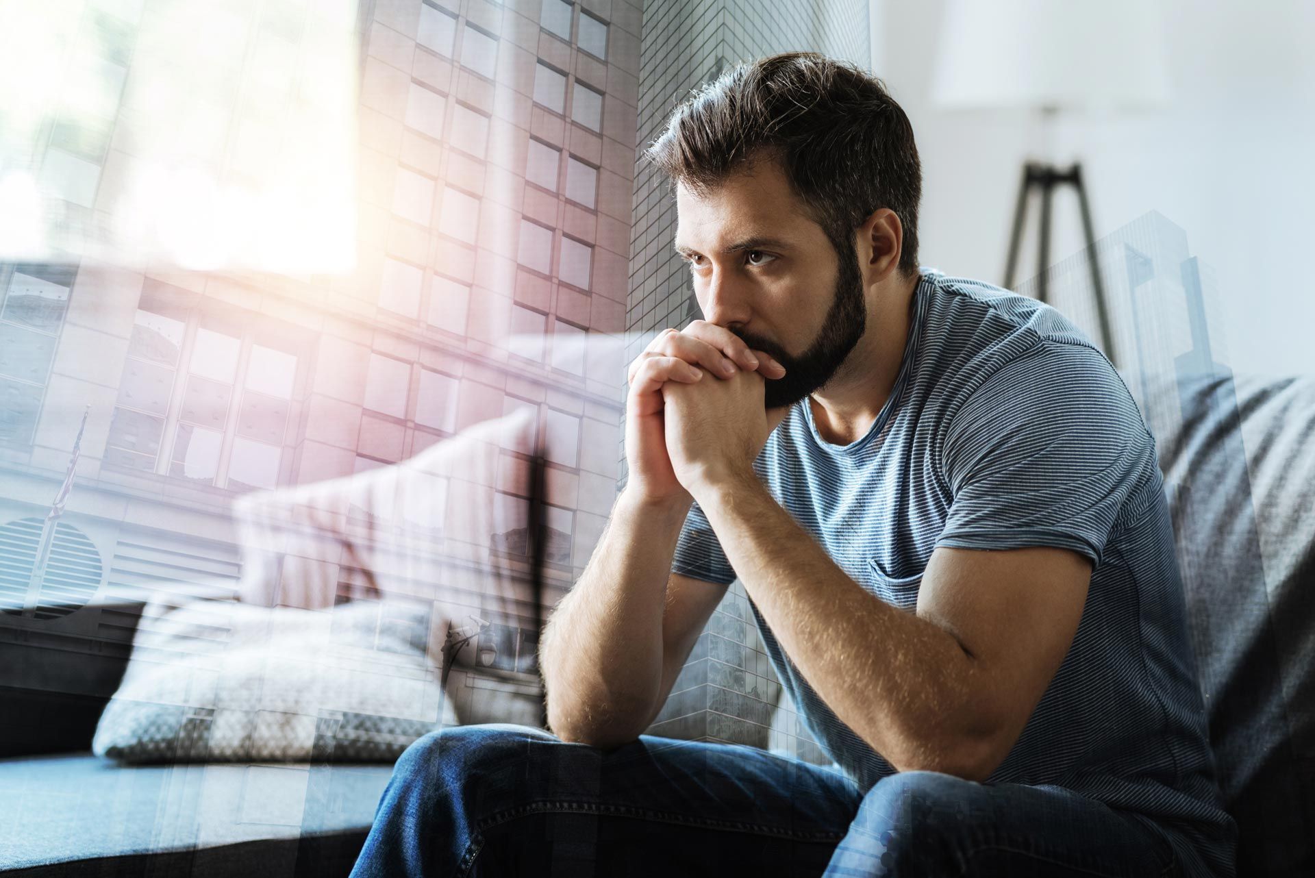 Man sitting on a couch with hands clasped, looking pensive in a bright living room