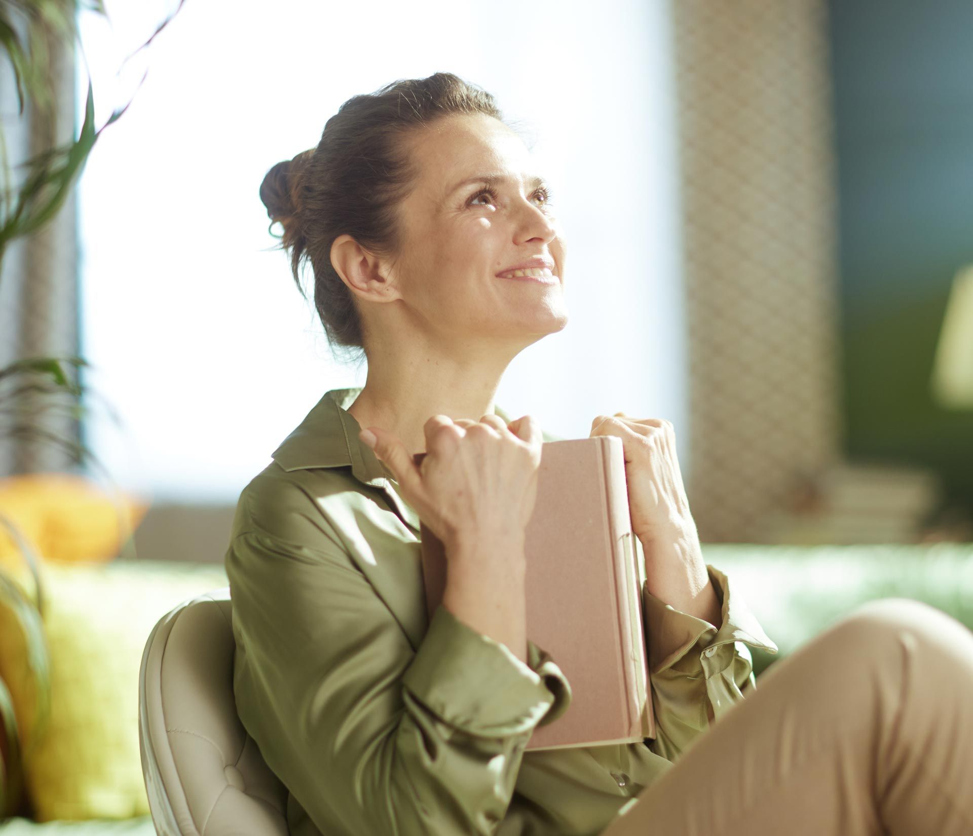 Smiling person holding a notebook in a bright room, looking upward thoughtfully.