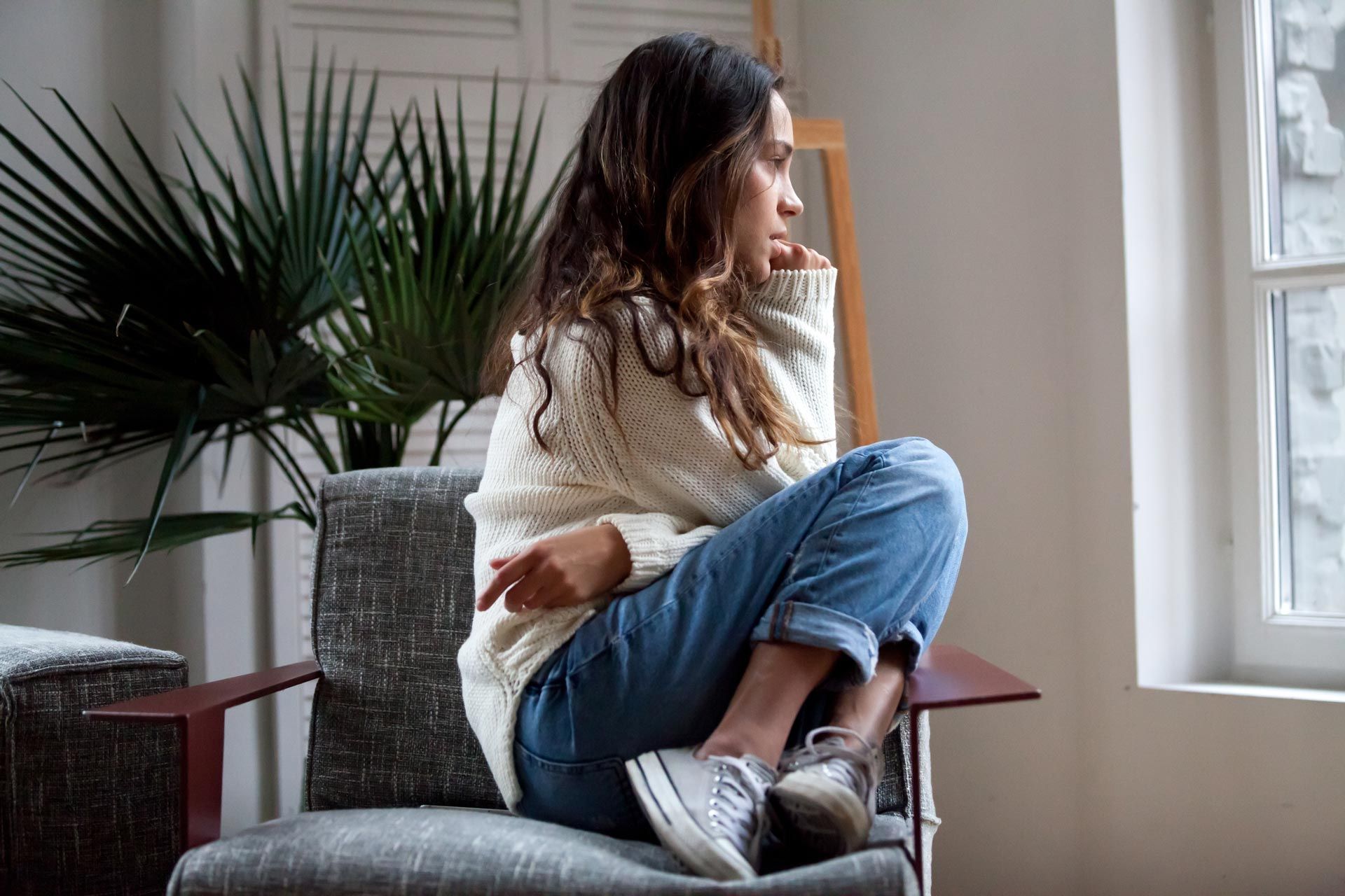 Woman sitting cross-legged on a chair by a window, looking outside, with a plant in the room.