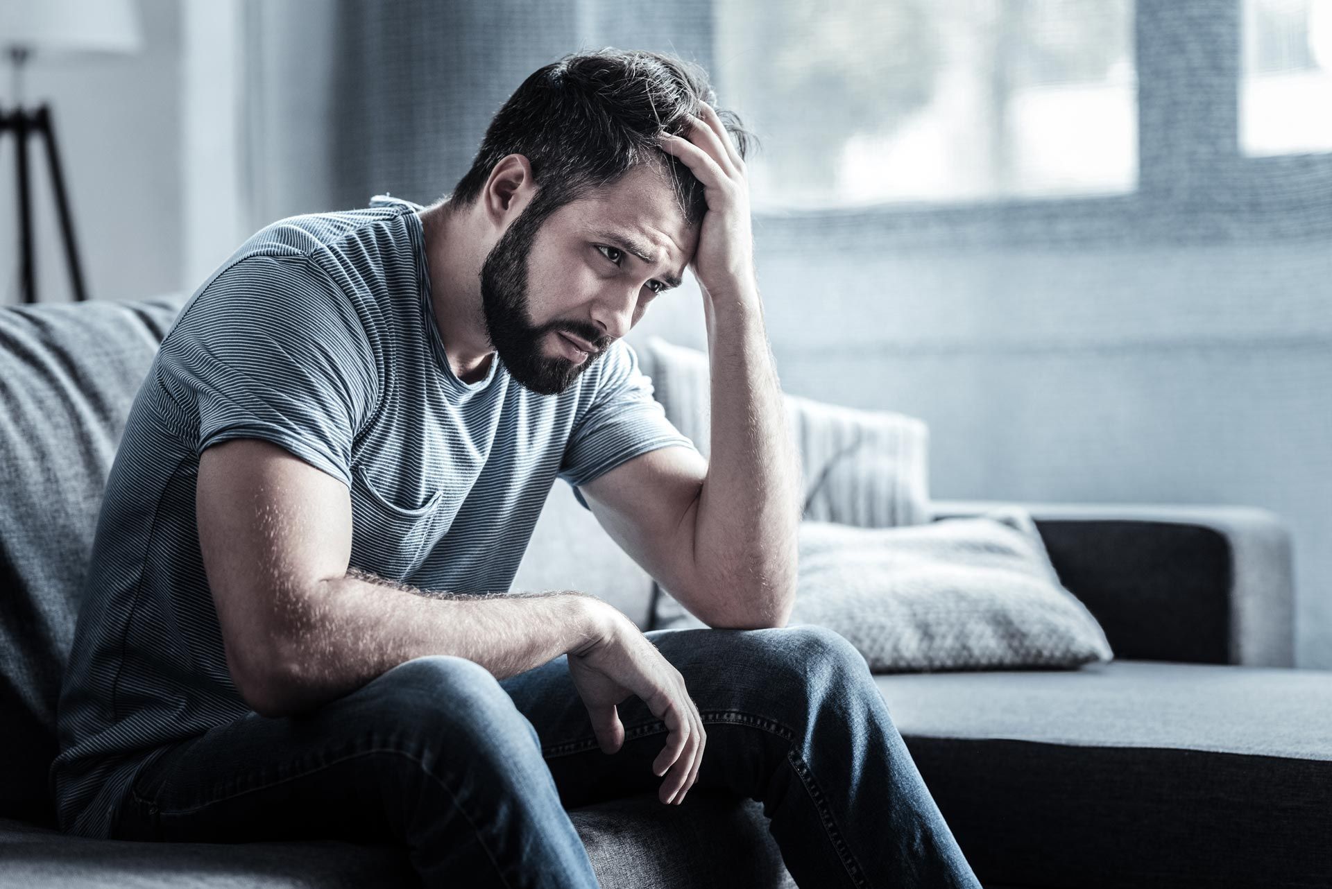 Man sitting on a couch, leaning forward with head in hand, appearing stressed in a dim living room