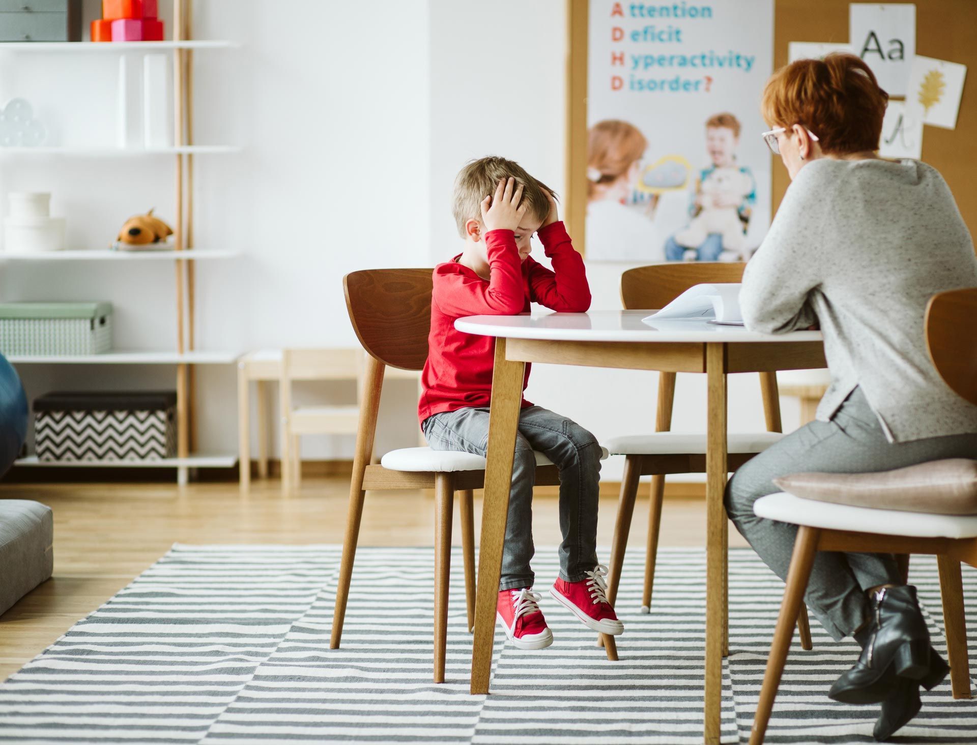 Child sitting at a table with an adult in a bright room, holding their head in their hands