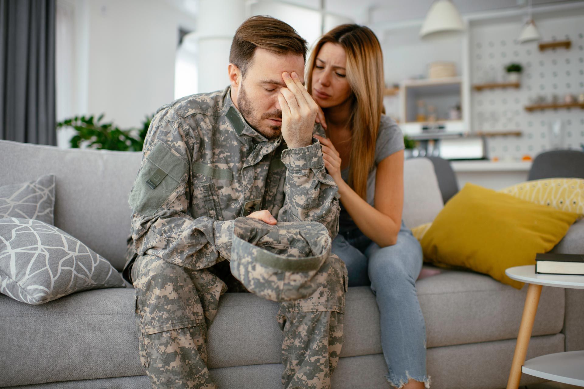 Soldier in camouflage sits on a couch, leaning forward while a woman comforts him in a living room