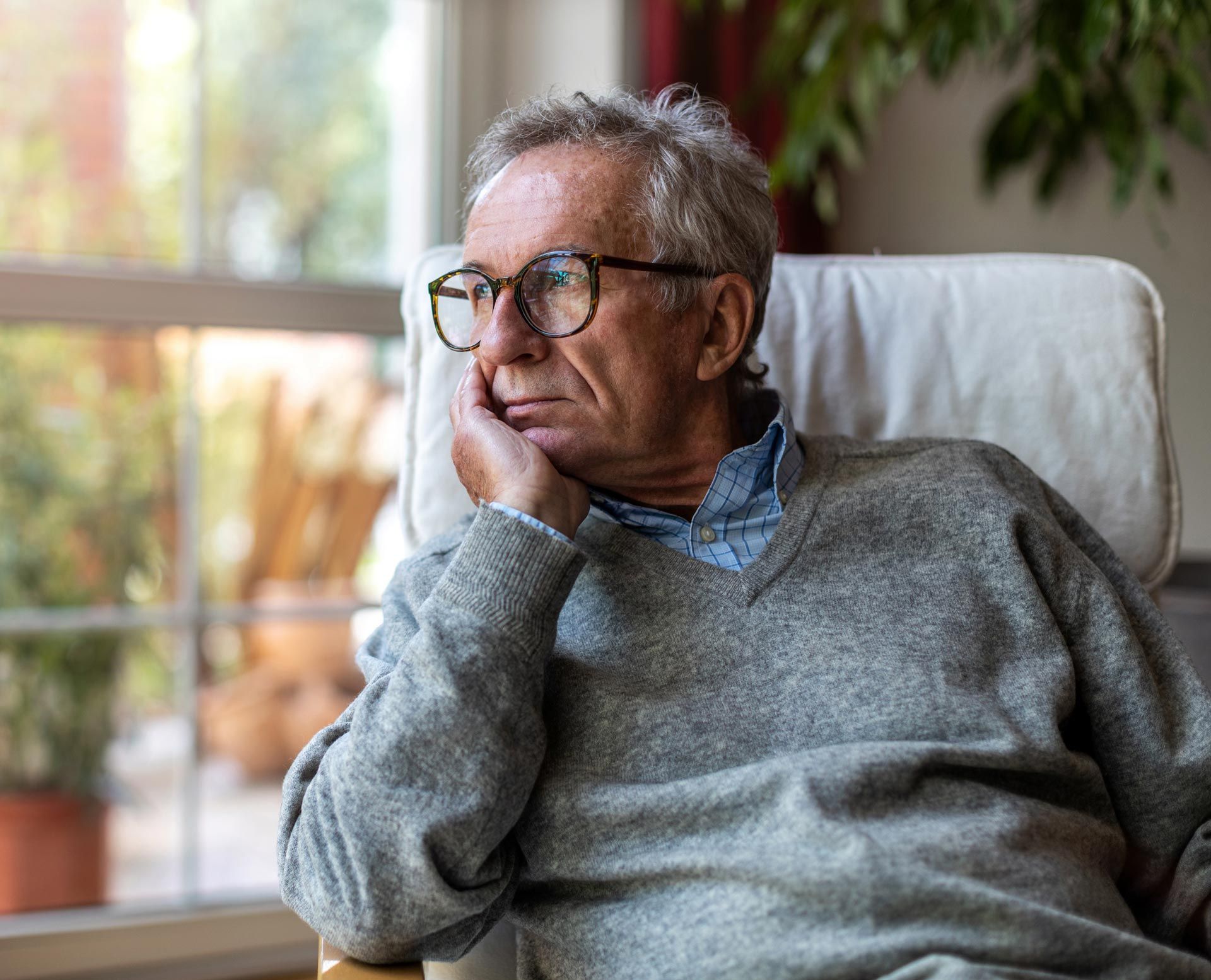 Man in glasses and gray sweater resting his head by a window, looking thoughtful.
