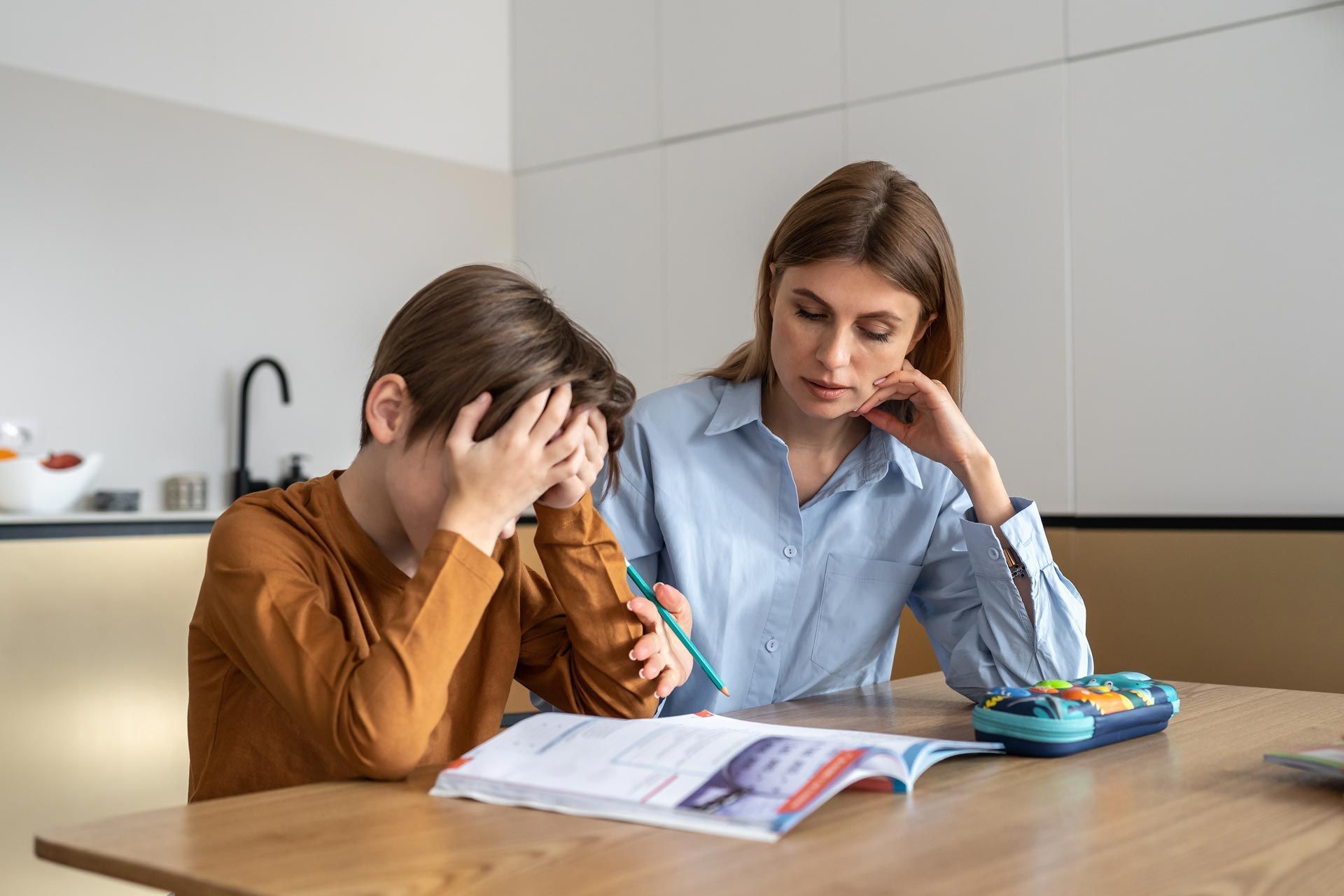 Two people studying at a kitchen table, looking frustrated over an open book and calculator.