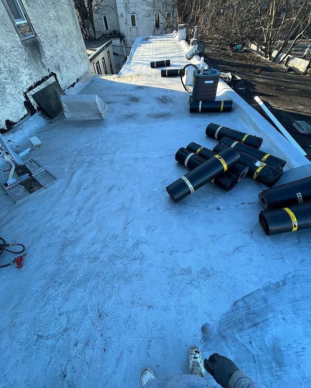 A person is standing on a roof with rolls of roofing material on it.