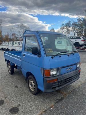 A light blue, small pickup truck parked on an asphalt lot under a cloudy sky.