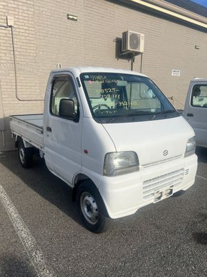 A white, compact single-cab kei truck parked on an asphalt lot in front of a light-colored brick building.