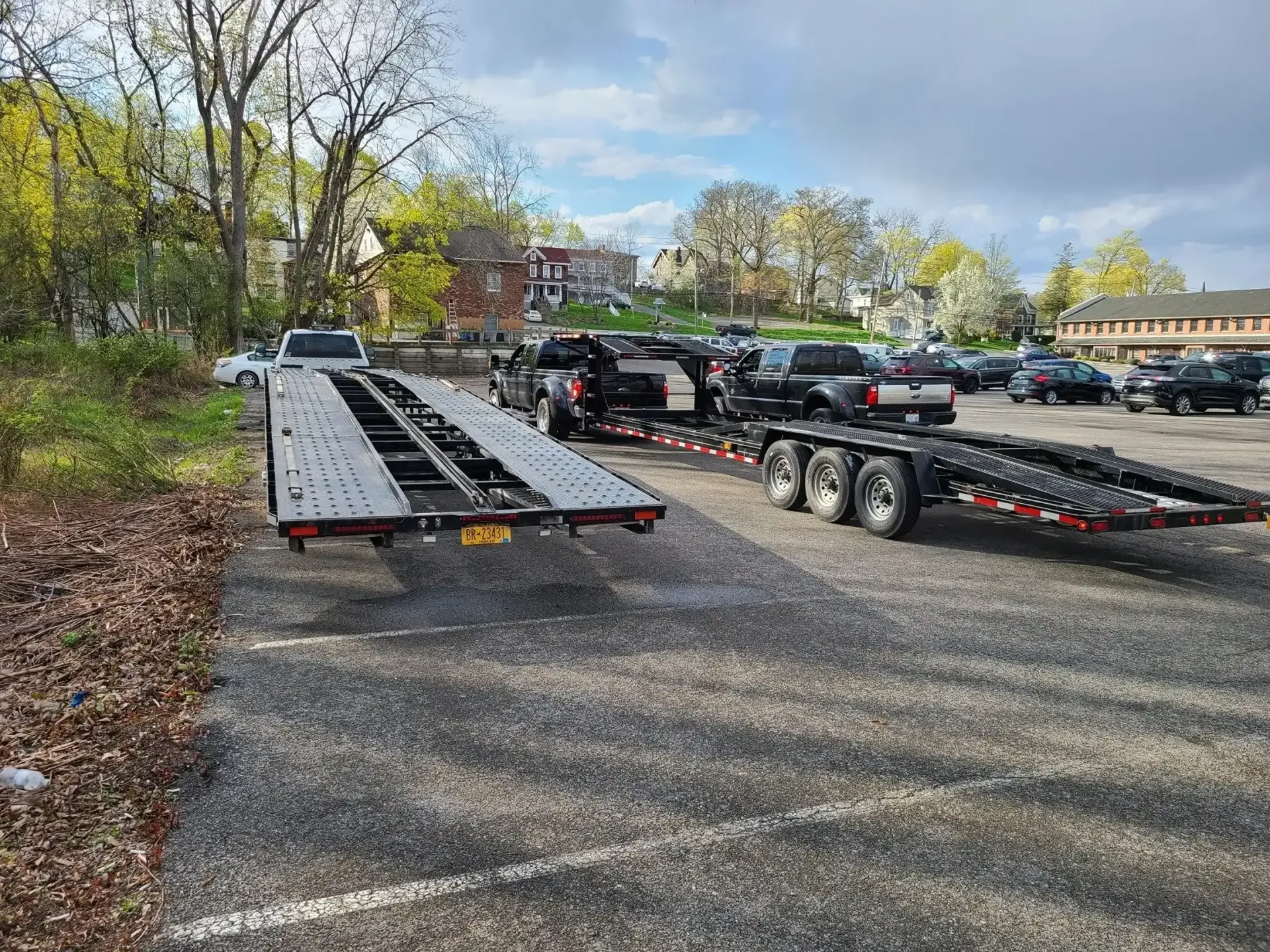 A large, empty flatbed car hauler trailer parked on a gravel lot next to trees and other vehicles.
