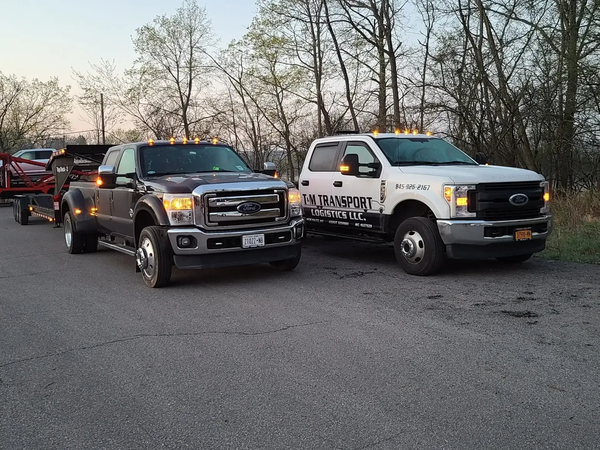 Two Ford F-series tow trucks parked side-by-side on a gravel road in front of a line of trees at sunset.