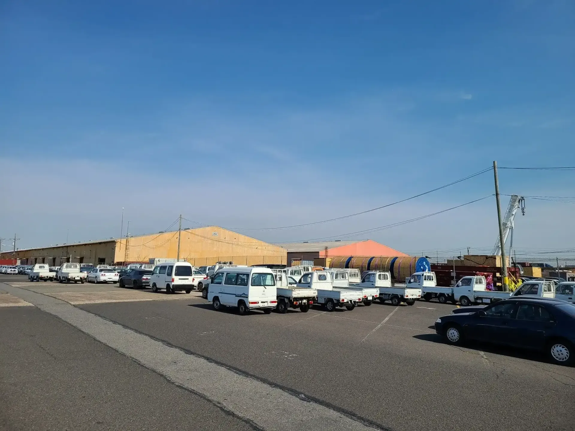 A gravel parking lot filled with various white work vans and trucks under a clear blue sky, next to large warehouses.