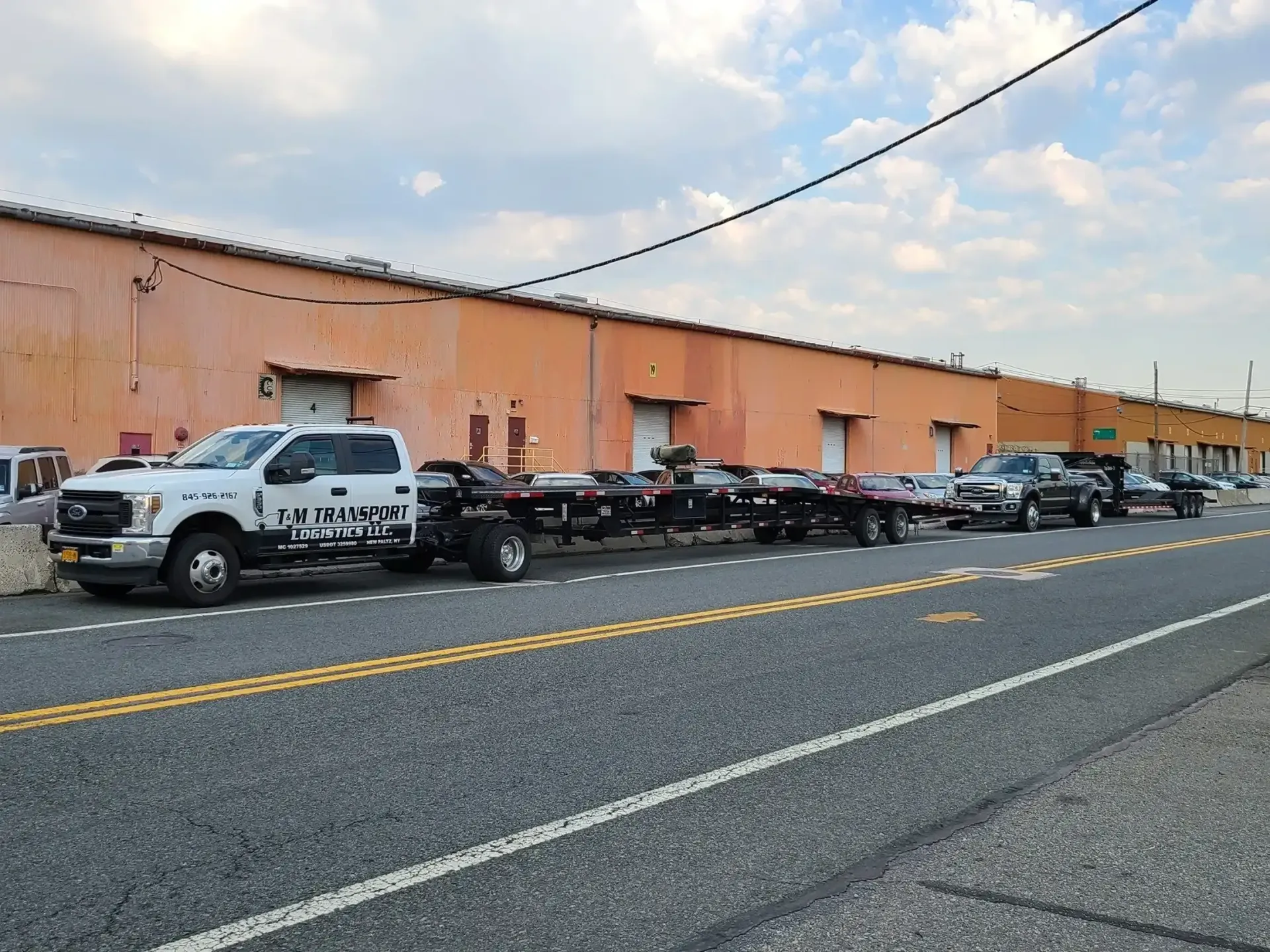 A white towing truck parked on the side of a road in front of a long, peach-colored industrial building.