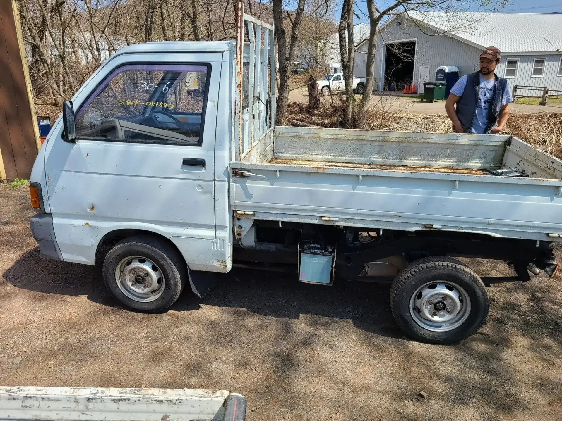 A light blue kei truck parked on a gravel surface outdoors with a person standing behind the bed.